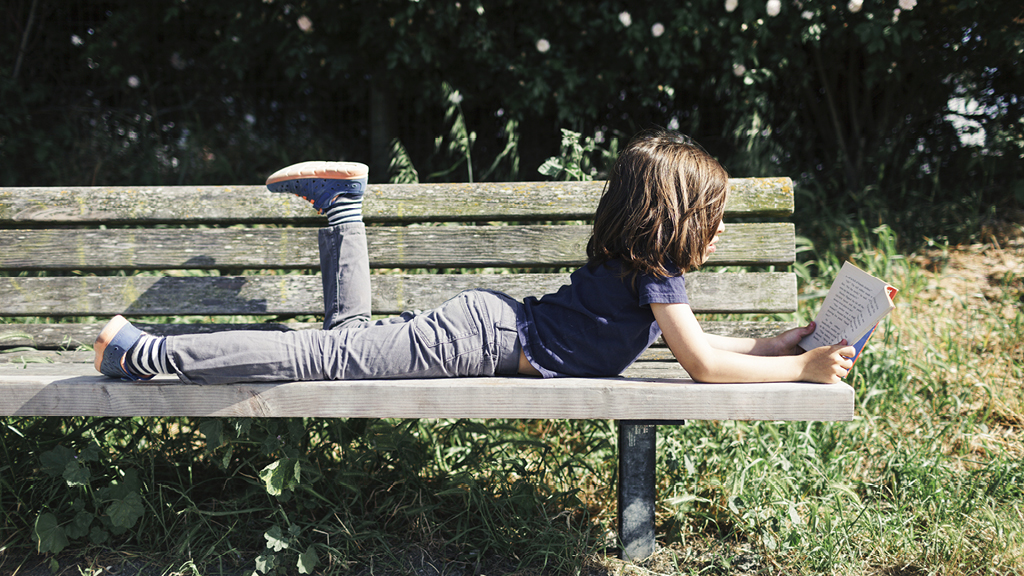 Child relaxing while reading outside on a bench.