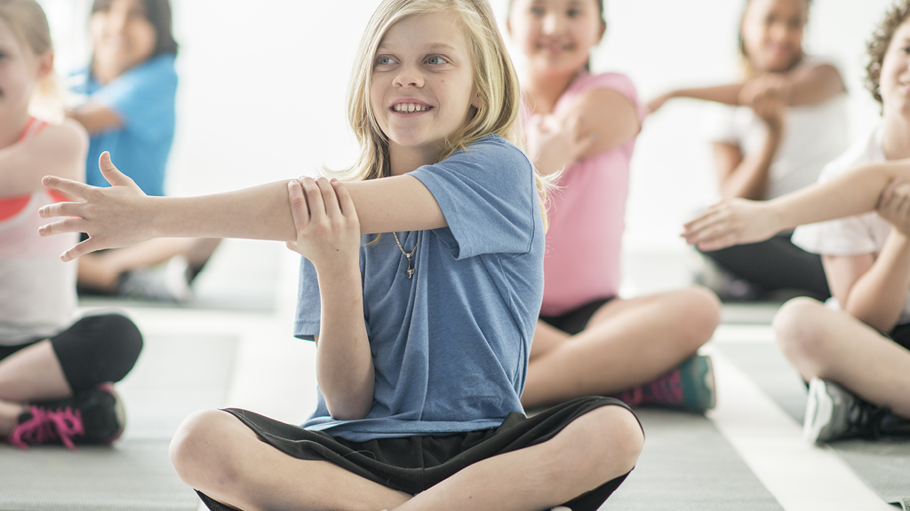 group of kids stretching while sitting cross-legged on a gym floor